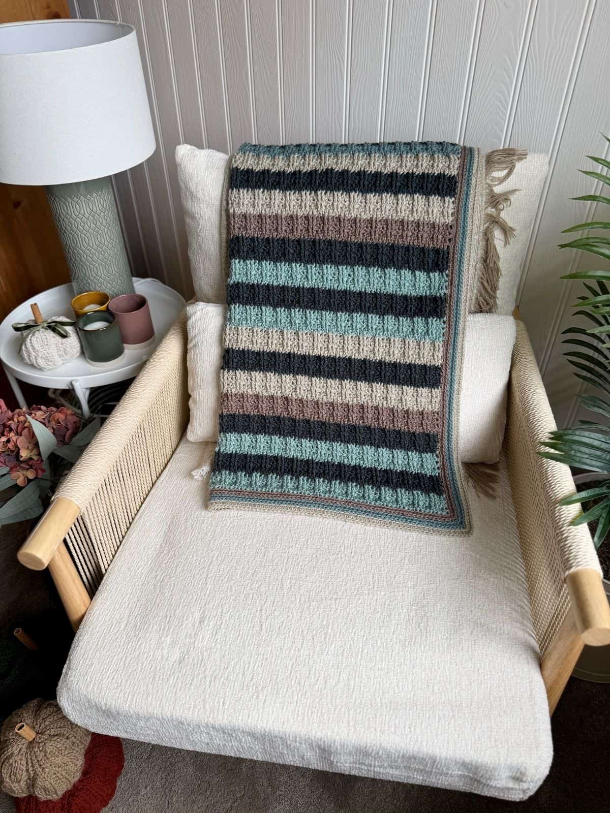 A striped crochet blanket for boys in earth tones is draped over a cream upholstered armchair in a cozy, decorated corner with a lamp, mugs, and plants nearby.