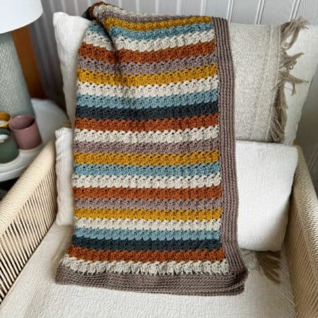 A crocheted blanket made using an easy crochet throw pattern, with brown, mustard yellow, gray, and rust-colored stripes, is draped over cream cushions on a beige chair.