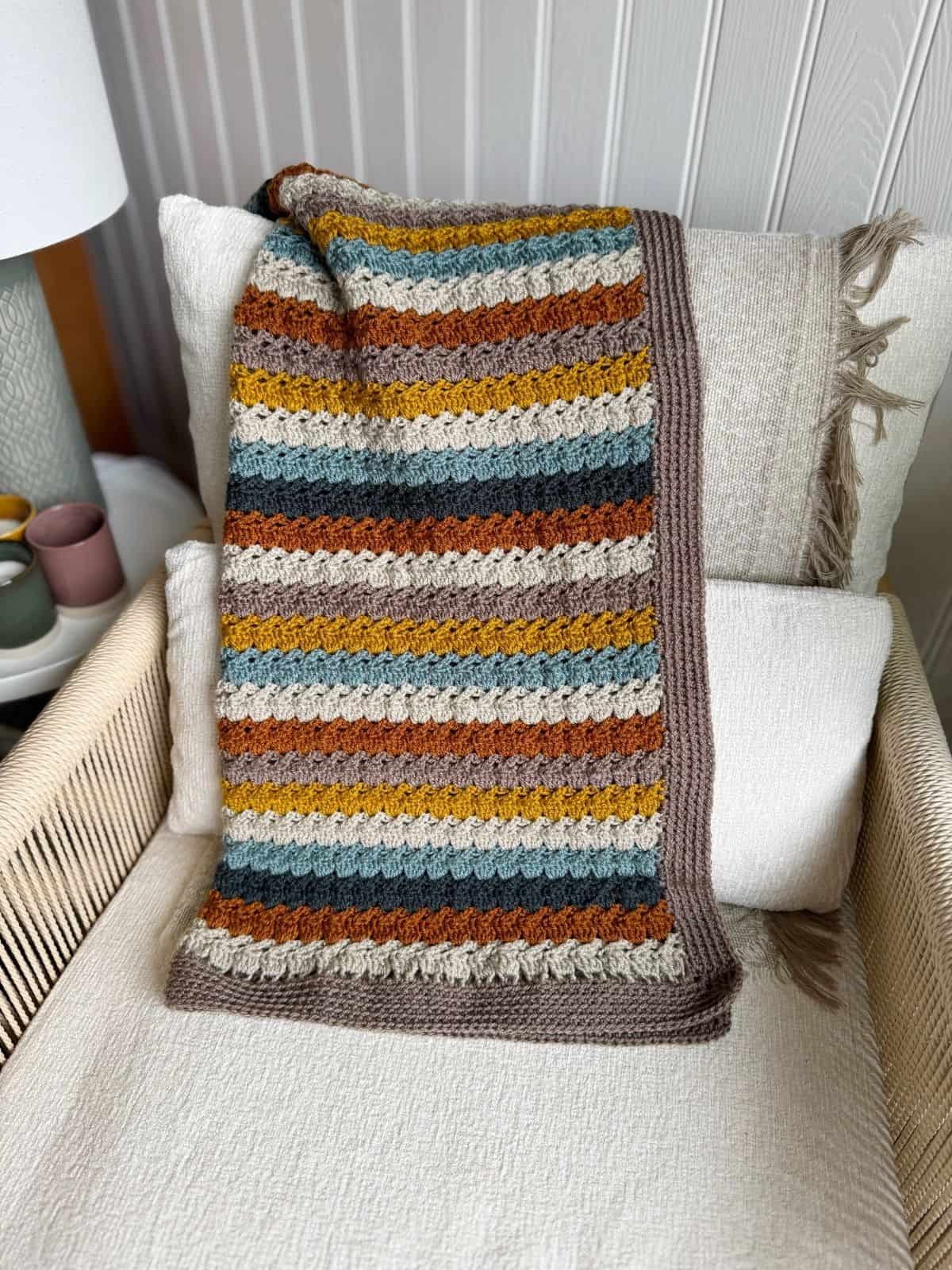 A crocheted blanket made using an easy crochet throw pattern, with brown, mustard yellow, gray, and rust-colored stripes, is draped over cream cushions on a beige chair.