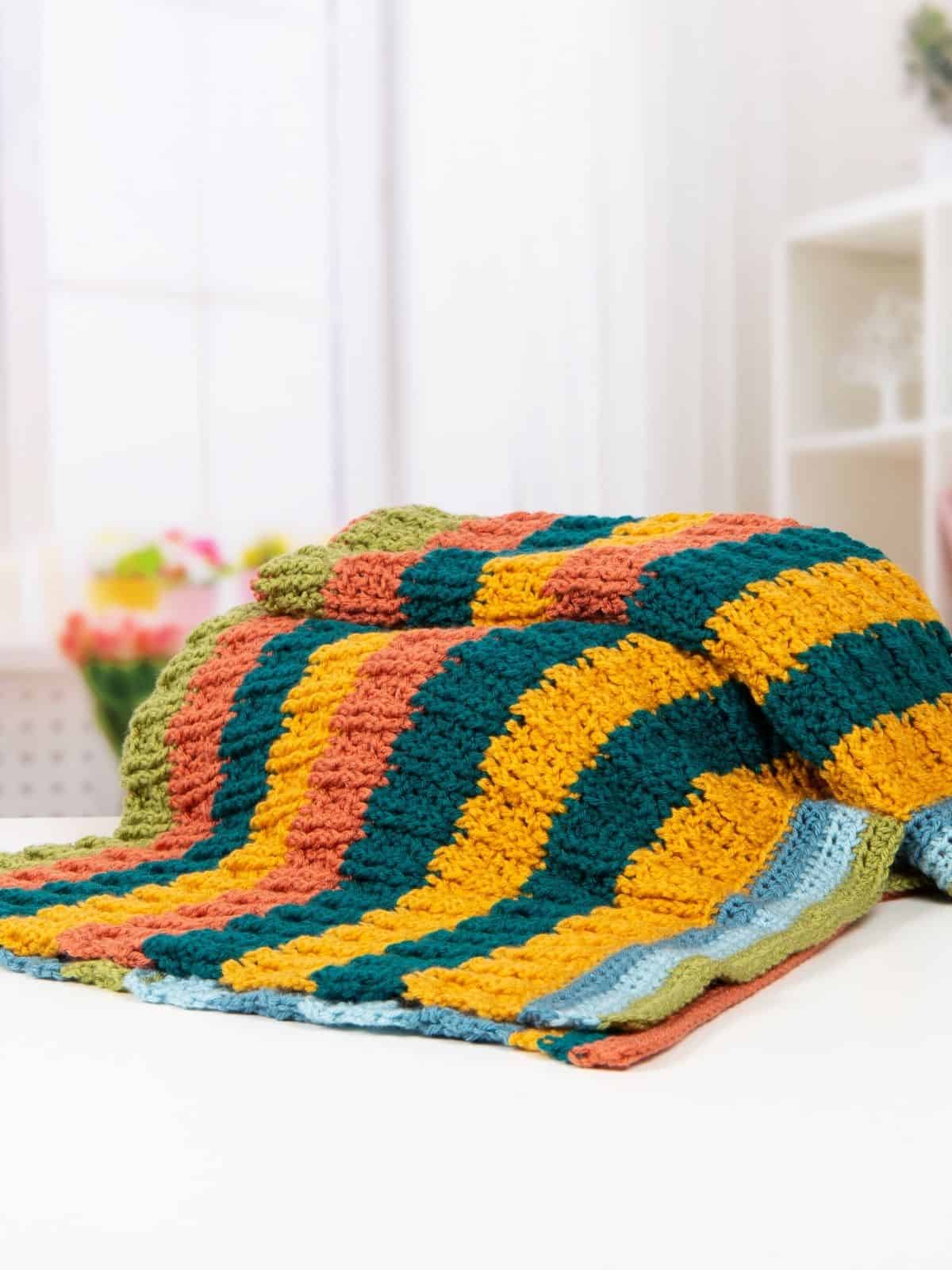 A colorful crochet blanket for boys with horizontal stripes in yellow, teal, orange, green, and light blue is draped over a white surface in a bright room.