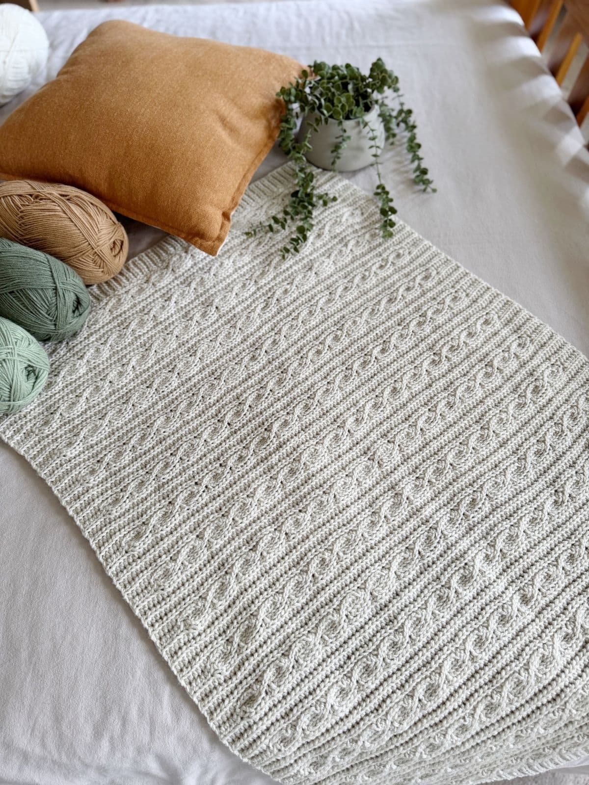 A textured, off-white crocheted blanket featuring a modern crochet blanket pattern is laid out on a bed next to balls of yarn, a brown cushion, and a small potted plant.
