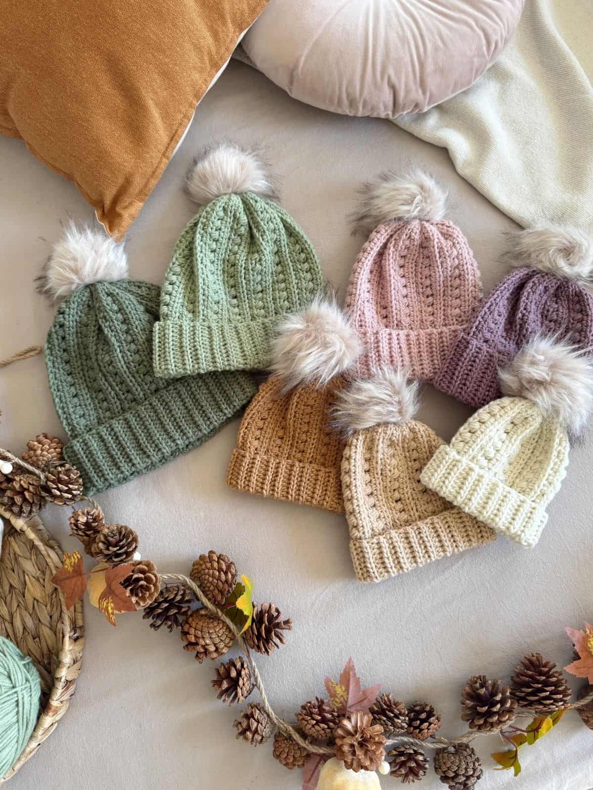Six pastel puff stitch crochet hats with faux fur pom-poms are arranged on a beige surface, surrounded by pine cones, yarn, and autumn leaves.