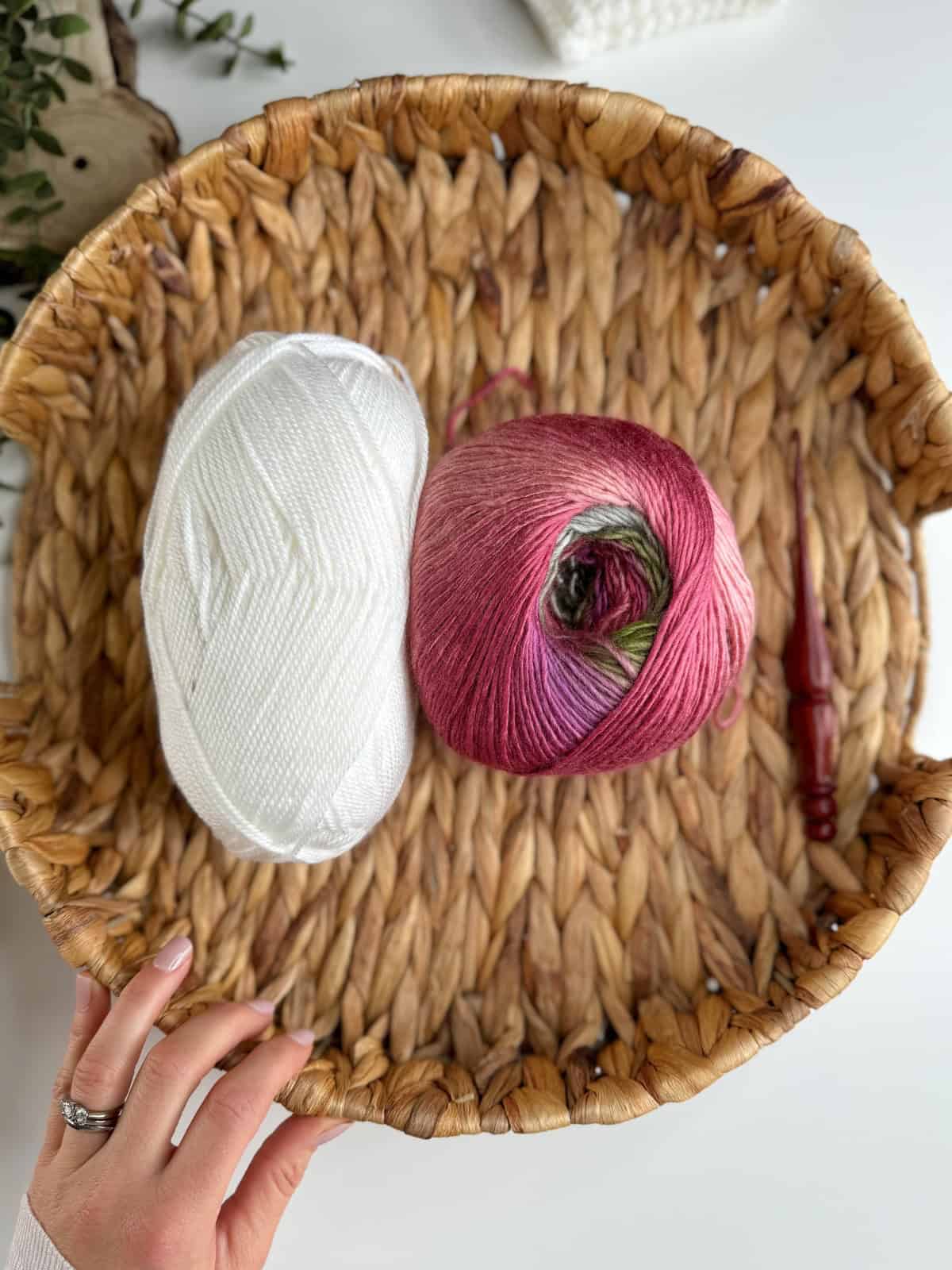 A hand holds a woven basket containing a skein of white yarn, a skein of variegated pink yarn, and a wooden crochet hook.