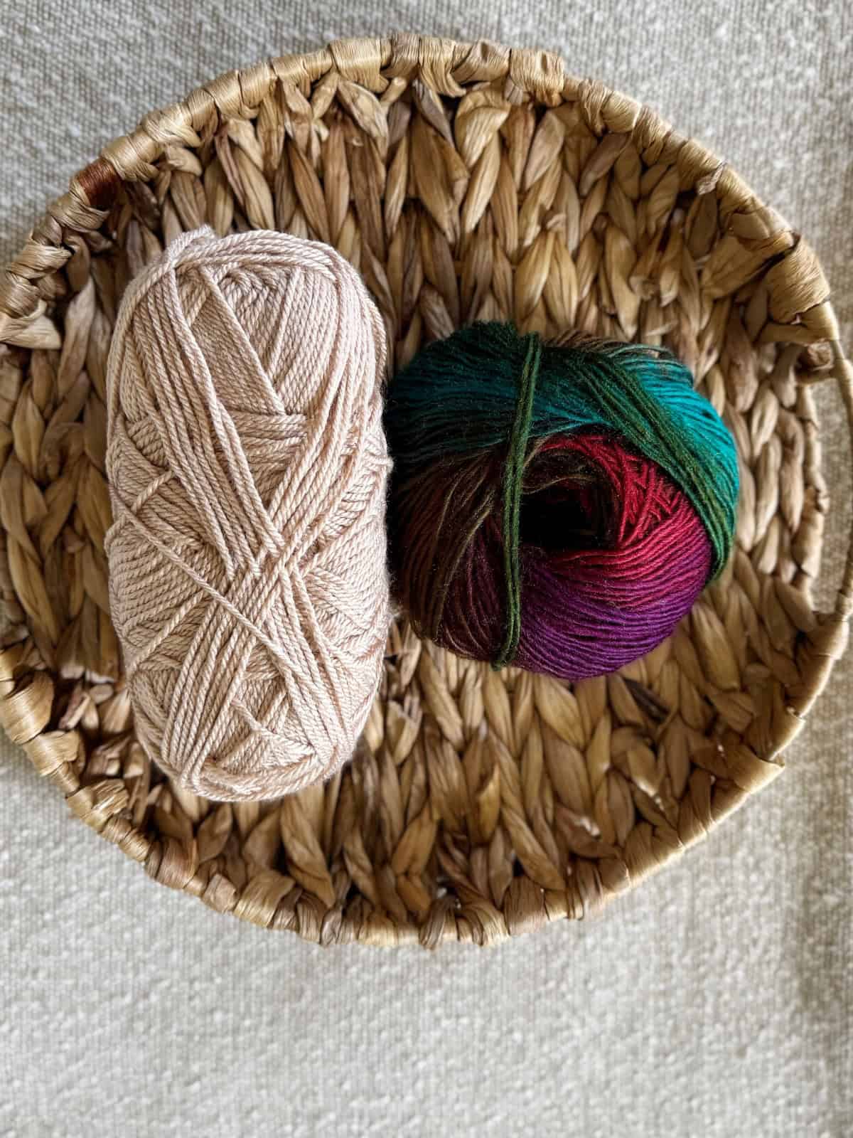 A woven basket holds two balls of yarn, one beige and one multicolored, on a textured light-colored surface.
