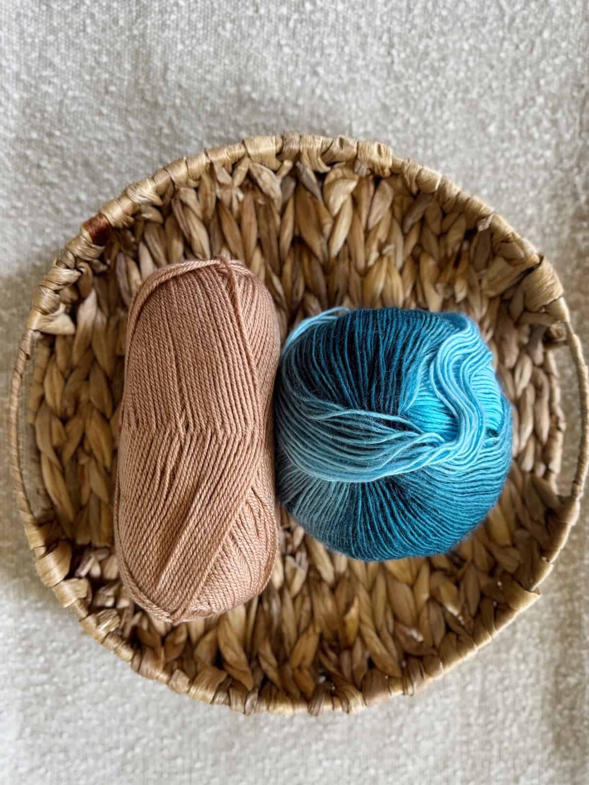 A round woven basket holds two skeins of yarn, one solid beige and one blue with gradient shades, placed on a textured light surface.