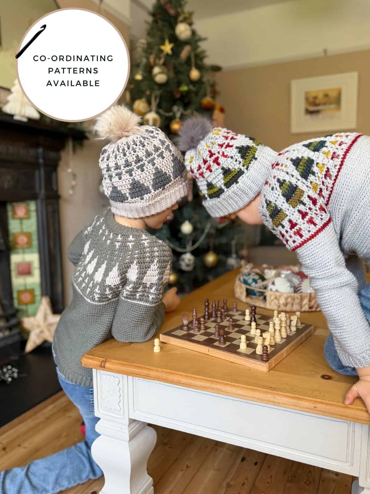 Two children wearing matching crochet sweaters and christmas tree crochet hat patterns play chess on a small table by a decorated Christmas tree. Text overlay reads “Co-ordinating patterns available.”.