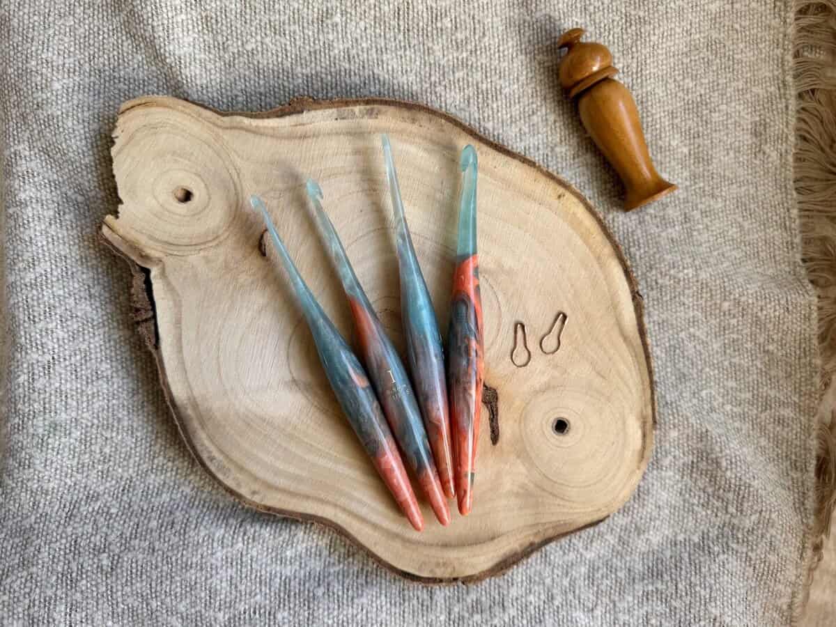 Four colorful crochet hooks in various crochet hook sizes and two stitch markers are displayed on a round wooden slab, with a wooden tool beside them, all placed on a textured beige fabric background.