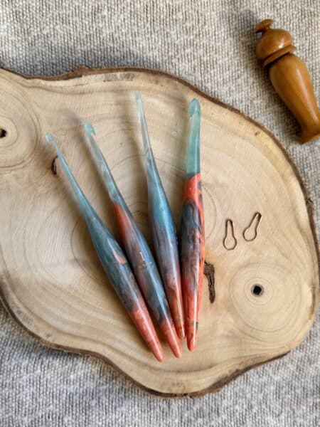 Four colorful crochet hooks in various crochet hook sizes and two stitch markers are displayed on a round wooden slab, with a wooden tool beside them, all placed on a textured beige fabric background.