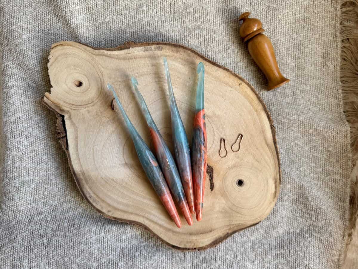Four colorful crochet hooks in various crochet hook sizes and two stitch markers are displayed on a round wooden slab, with a wooden tool beside them, all placed on a textured beige fabric background.