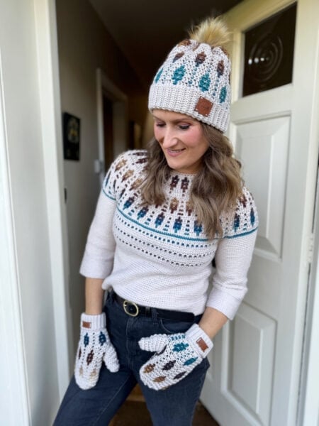 Woman wearing a crocheted hat, sweater, and mittens with a matching pattern of brown and teal designs, standing indoors near a white door.