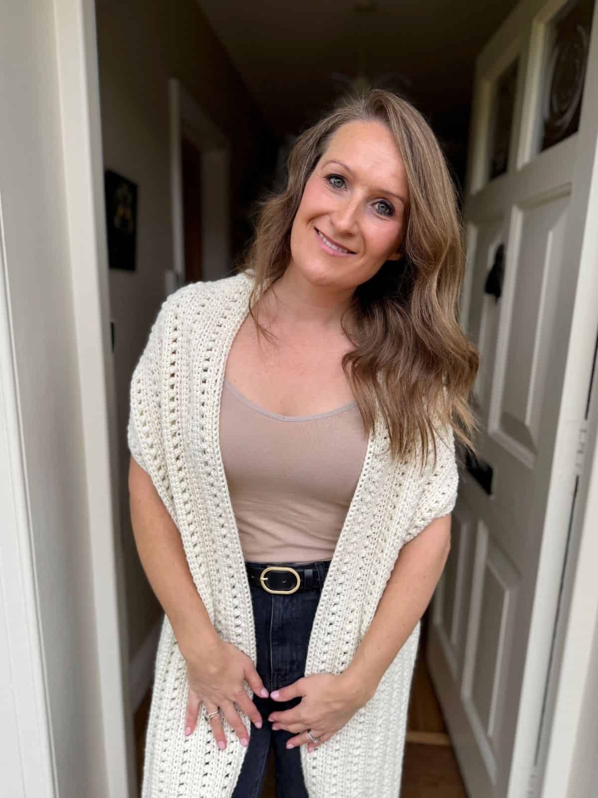 Woman standing in a doorway, wearing a beige top, black pants with a belt, and a white cardigan styled like a crochet sweater—get inspired by this look for your next crochet sweater scarf pattern free project.