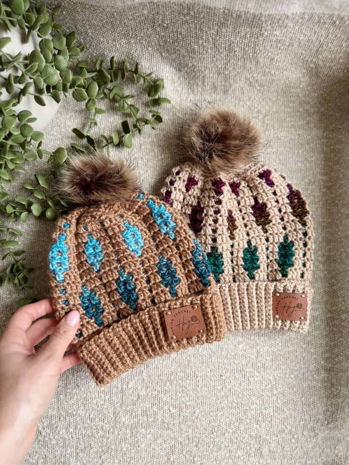 Two crocheted beanies with faux fur pom-poms and colorful diamond patterns are displayed on a textured fabric near a green plant. A hand holds the beanie on the left.