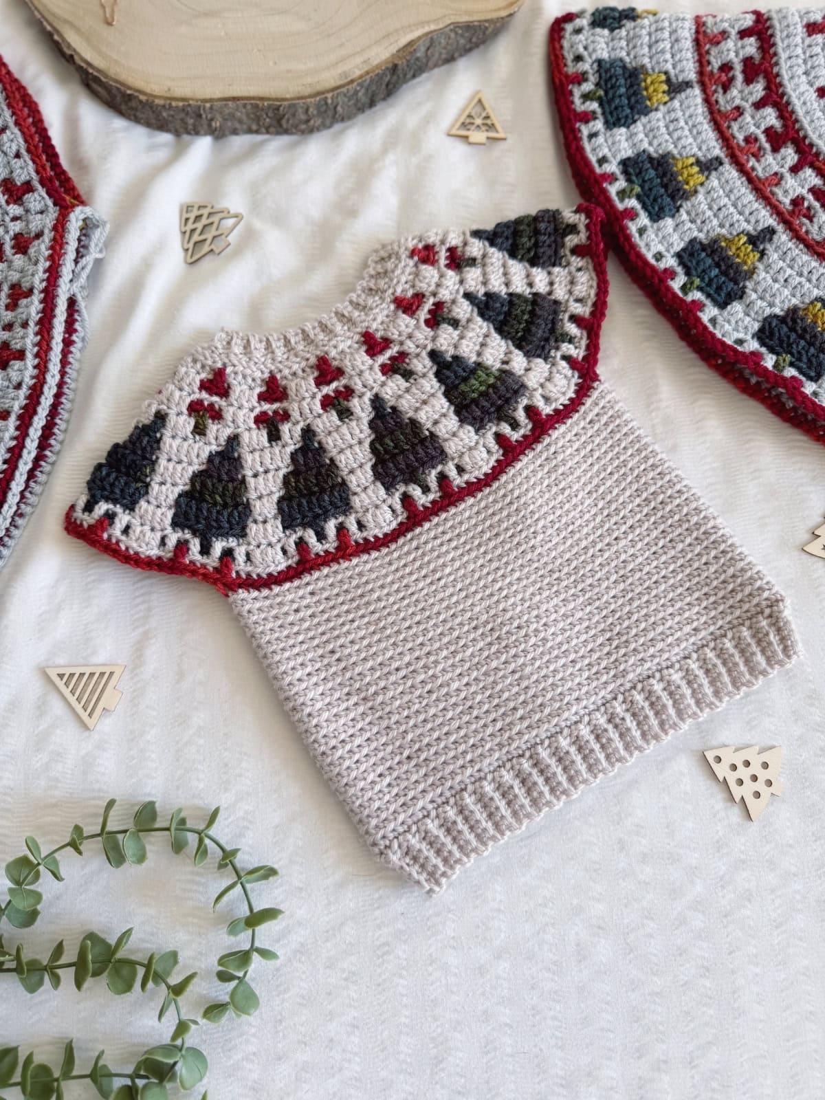 Hand-crocheted baby sweater with a patterned yoke in red, green, gray, and black—perfect as a crochet Christmas sweater—laid flat on a white textured surface with wooden decorations.
