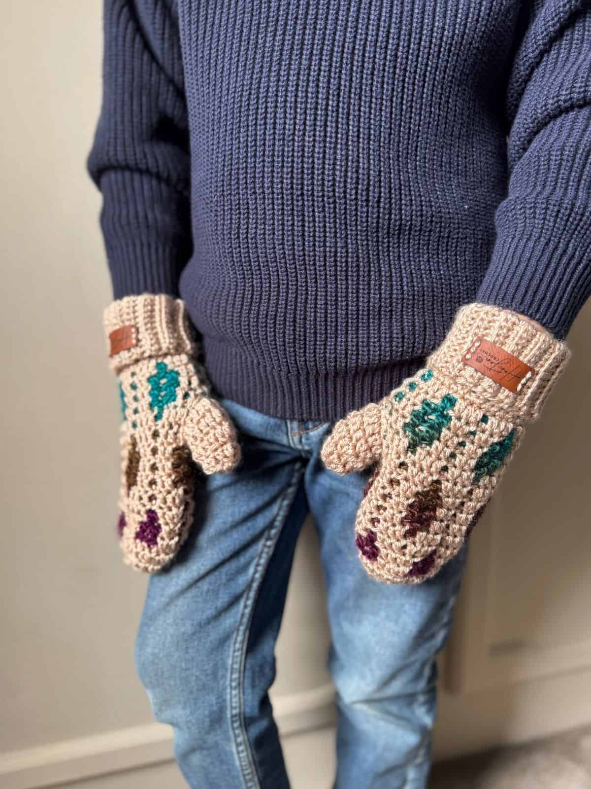 Person wearing a navy sweater, blue jeans, and beige mittens featuring a mosaic crochet mittens pattern with colorful accents stands with hands by their sides.