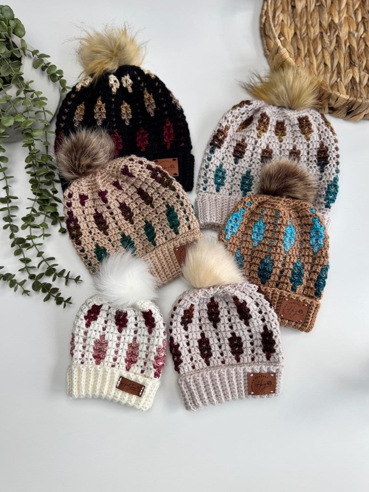 Six crocheted beanies with leaf patterns and faux fur pom-poms, including a stylish mosaic crochet beanie, are arranged on a white surface with greenery and a woven tray nearby.