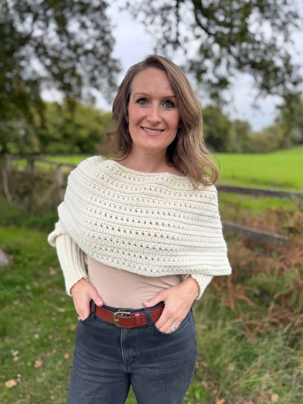 A woman stands outdoors in front of a wooden fence, wearing a cream-colored crochet sweater scarf, beige top, and dark jeans, with greenery and trees in the background—perfect inspiration for your next free pattern.
