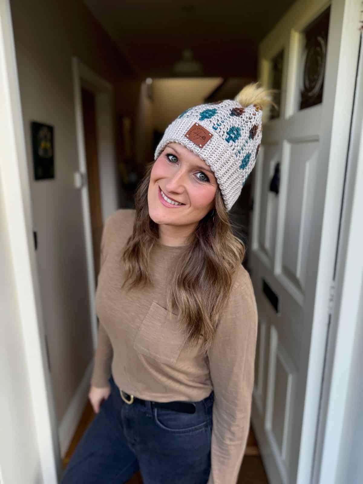 Woman stands in a doorway, smiling at the camera, wearing a beige long-sleeve shirt, black jeans, and a mosaic crochet beanie with a pom-pom.