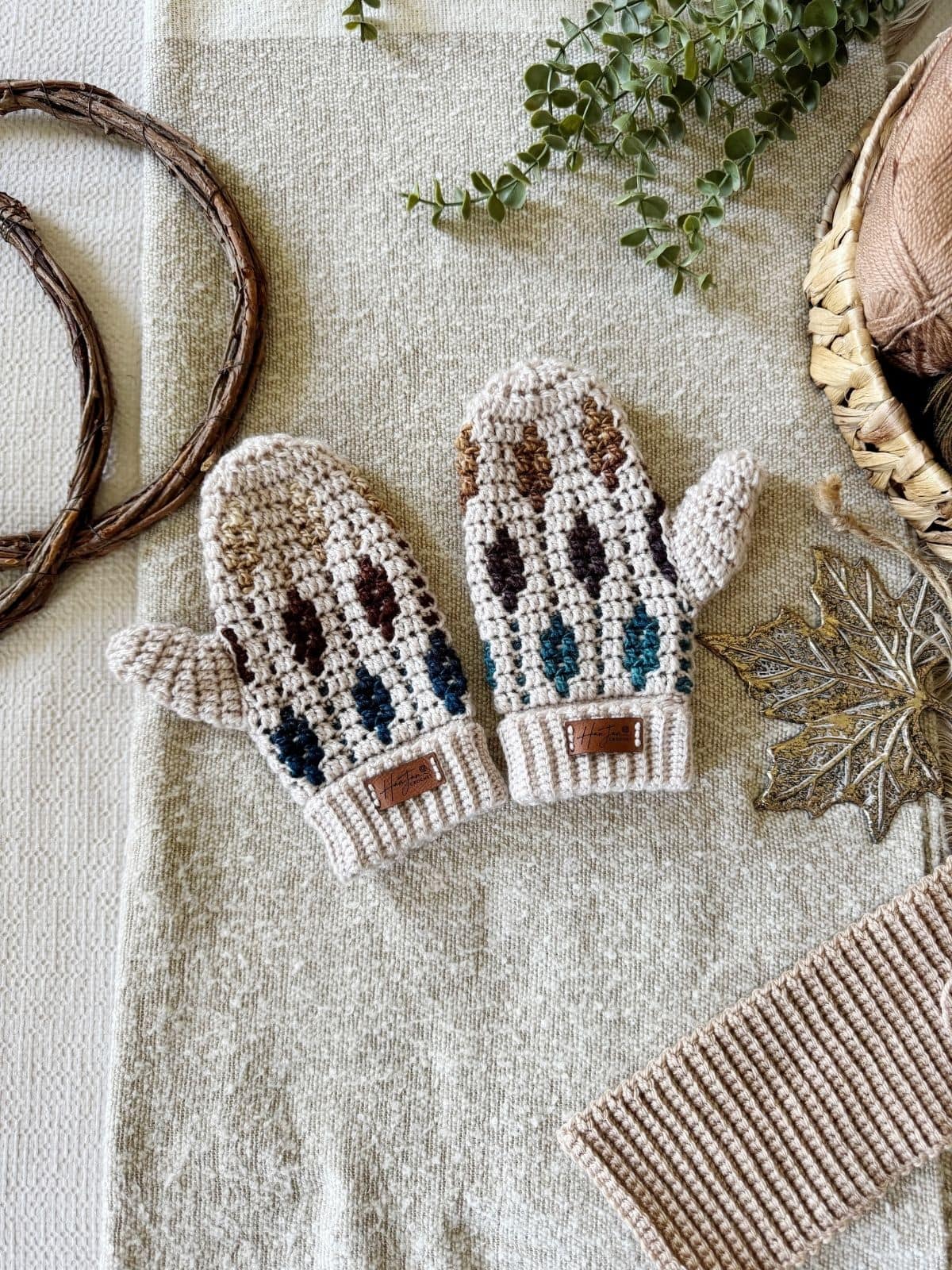 Two crocheted mittens featuring a vibrant mosaic crochet mittens pattern rest on textured fabric, surrounded by autumn-themed decor like a metal leaf, twigs, and greenery.