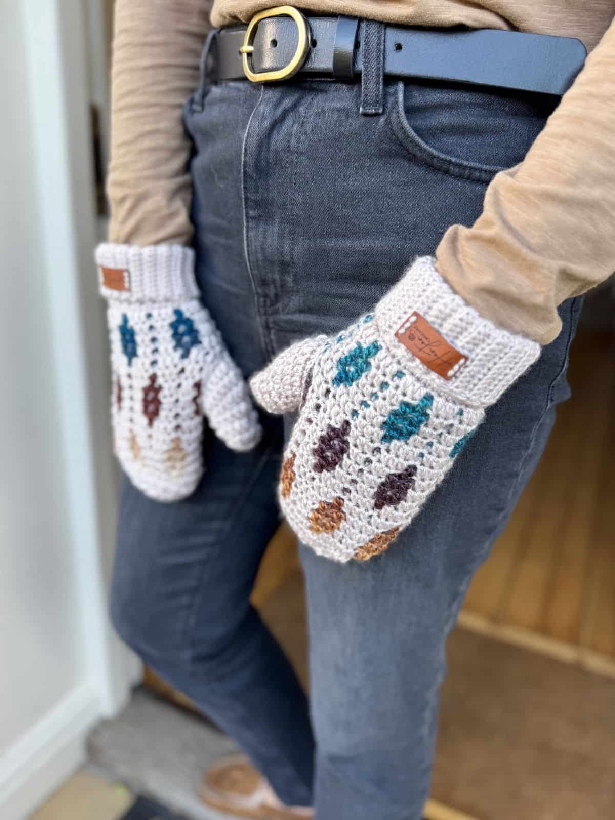 Person wearing gray jeans, a tan long-sleeve shirt, and a black belt, standing indoors with hands in front, displaying crochet mittens with multicolored leaf patterns inspired by a mosaic crochet mittens pattern.