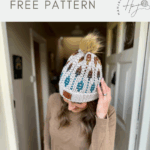 A woman wearing a crocheted hat with a pom-pom stands indoors beneath a sign reading "crochet hat pattern FREE PATTERN HanJan Crochet," showcasing her stylish mosaic crochet beanie.