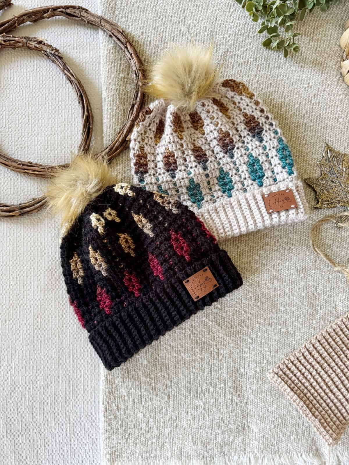 Two crochet beanies with faux fur pom-poms—one black with red and yellow accents, one cream with teal and brown accents—are displayed on a neutral woven surface. The mosaic crochet beanie design adds unique texture and style.