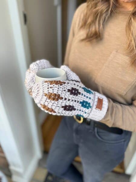 A person wearing a beige long-sleeve shirt and mosaic crochet mittens holds a gray mug while standing near a doorway.