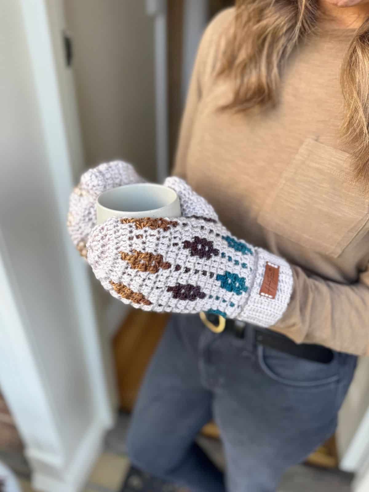 A person wearing a beige long-sleeve shirt and mosaic crochet mittens holds a gray mug while standing near a doorway.