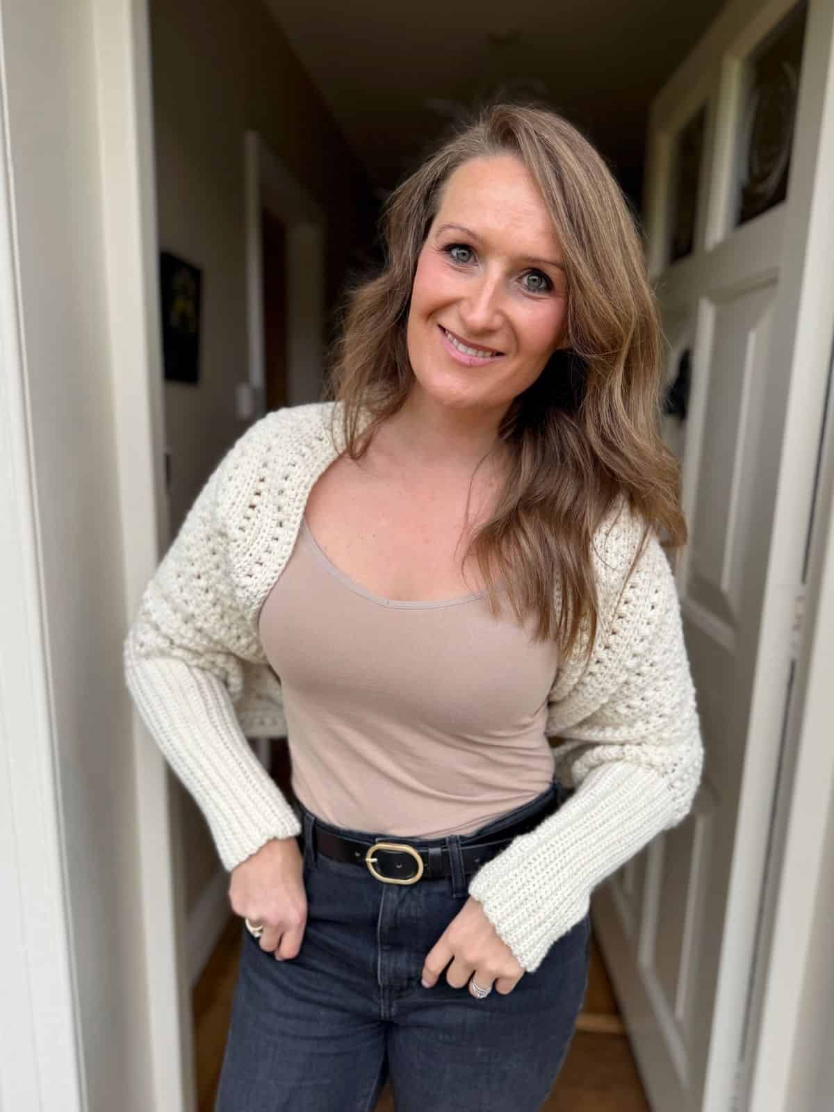 A woman with long brown hair stands in a doorway, smiling at the camera in a cream crochet cardigan layered over a beige top and dark jeans, accessorized with a crochet sweater scarf pattern free for cozy style.