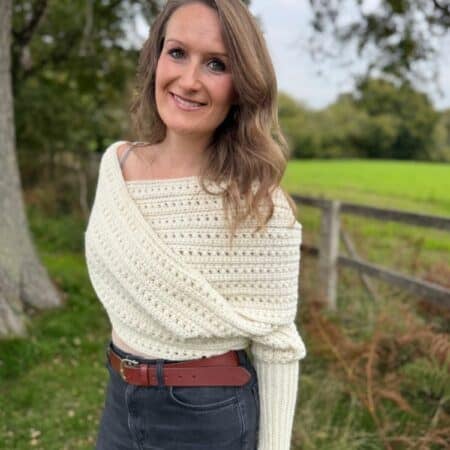 Woman standing outdoors in front of a wooden fence and grassy field, wearing a cream knit sweater and dark jeans, smiling at the camera.