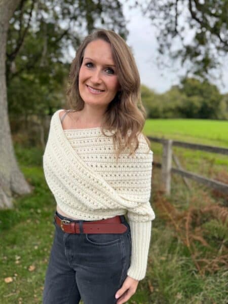 Woman standing outdoors in front of a wooden fence and grassy field, wearing a cream knit sweater and dark jeans, smiling at the camera.