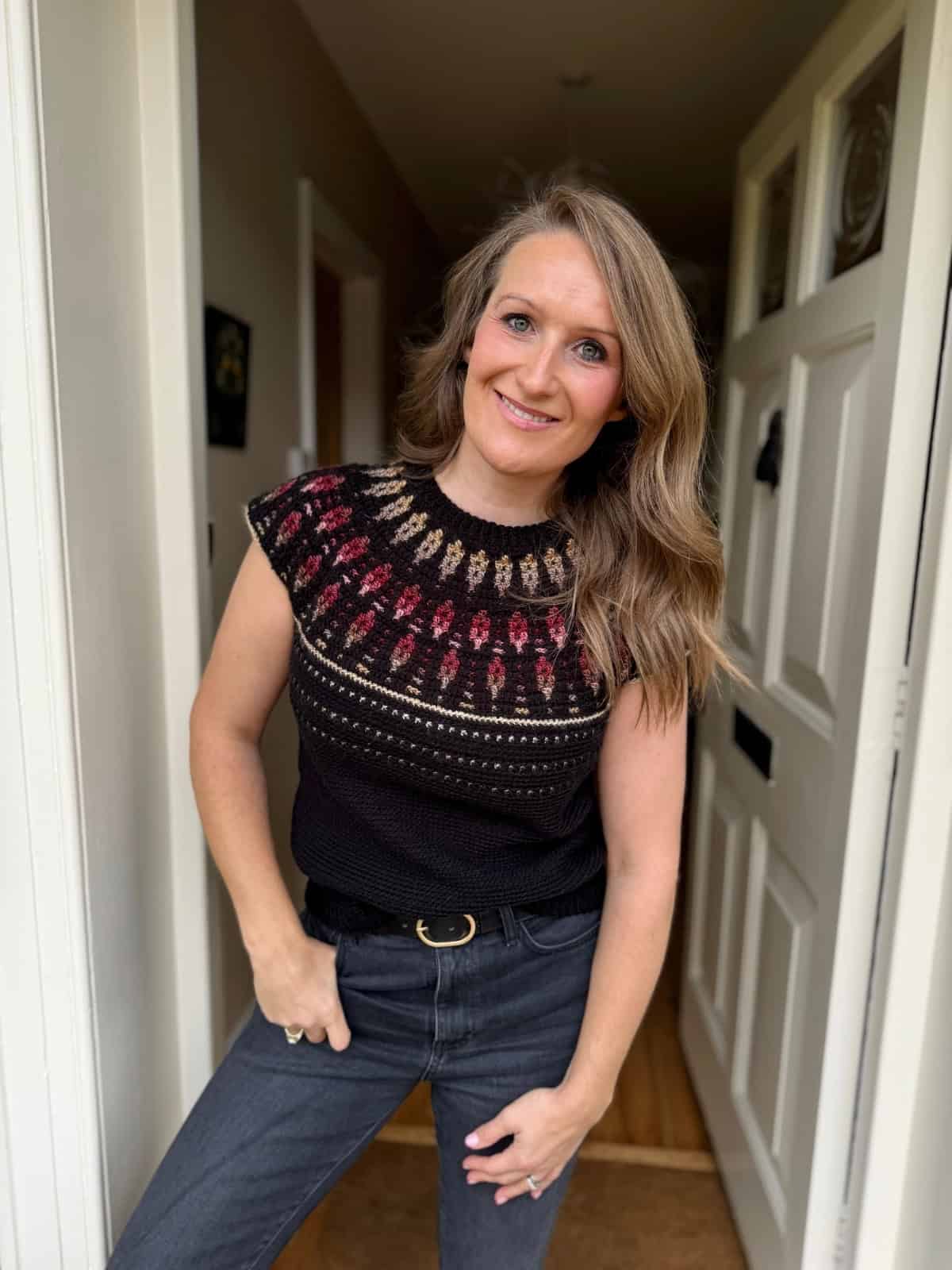 A woman stands in a doorway, smiling at the camera in a black knitted top featuring a mosaic crochet sweater pattern on the yoke, paired with dark jeans.