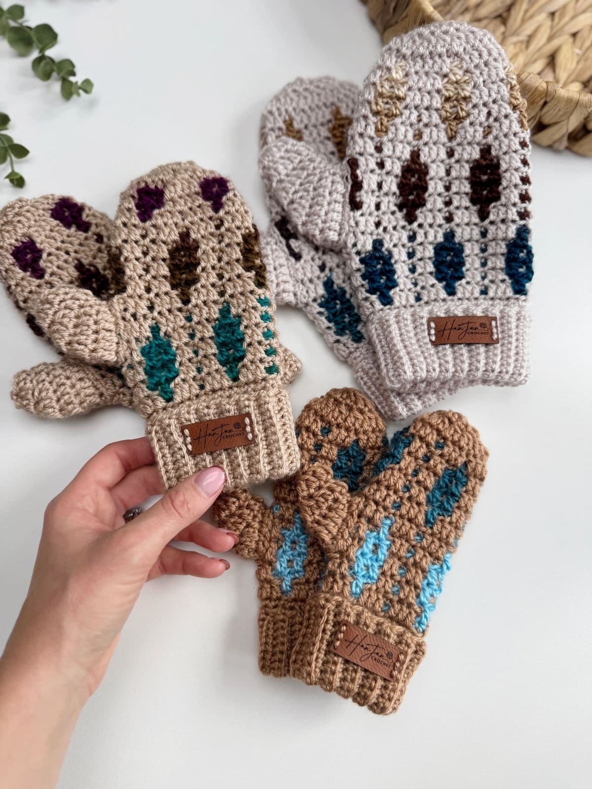 Three pairs of crocheted mittens with leaf designs, made using a mosaic crochet mittens pattern in different colors, are displayed on a white surface; a hand holds one mitten in the foreground.
