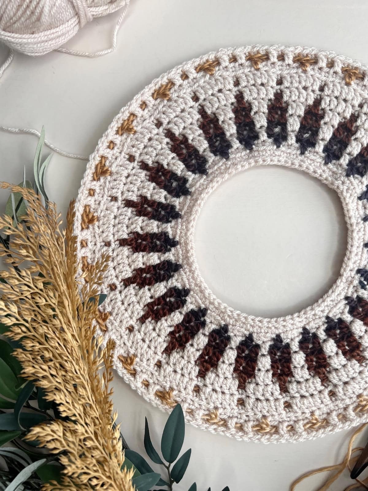 A round, crocheted piece with brown and tan patterns—reminiscent of a mosaic crochet sweater pattern—sits on a white surface beside a ball of yarn and some dried plants with green leaves.