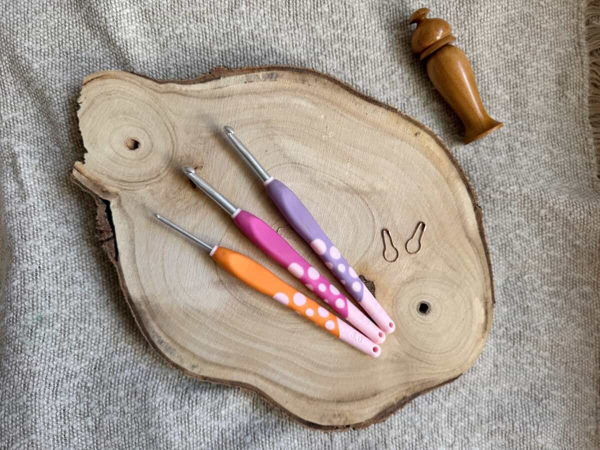 Three crochet hooks in assorted crochet hook sizes with pink and orange handles, two stitch markers, and a wooden container are arranged on a circular wooden board.