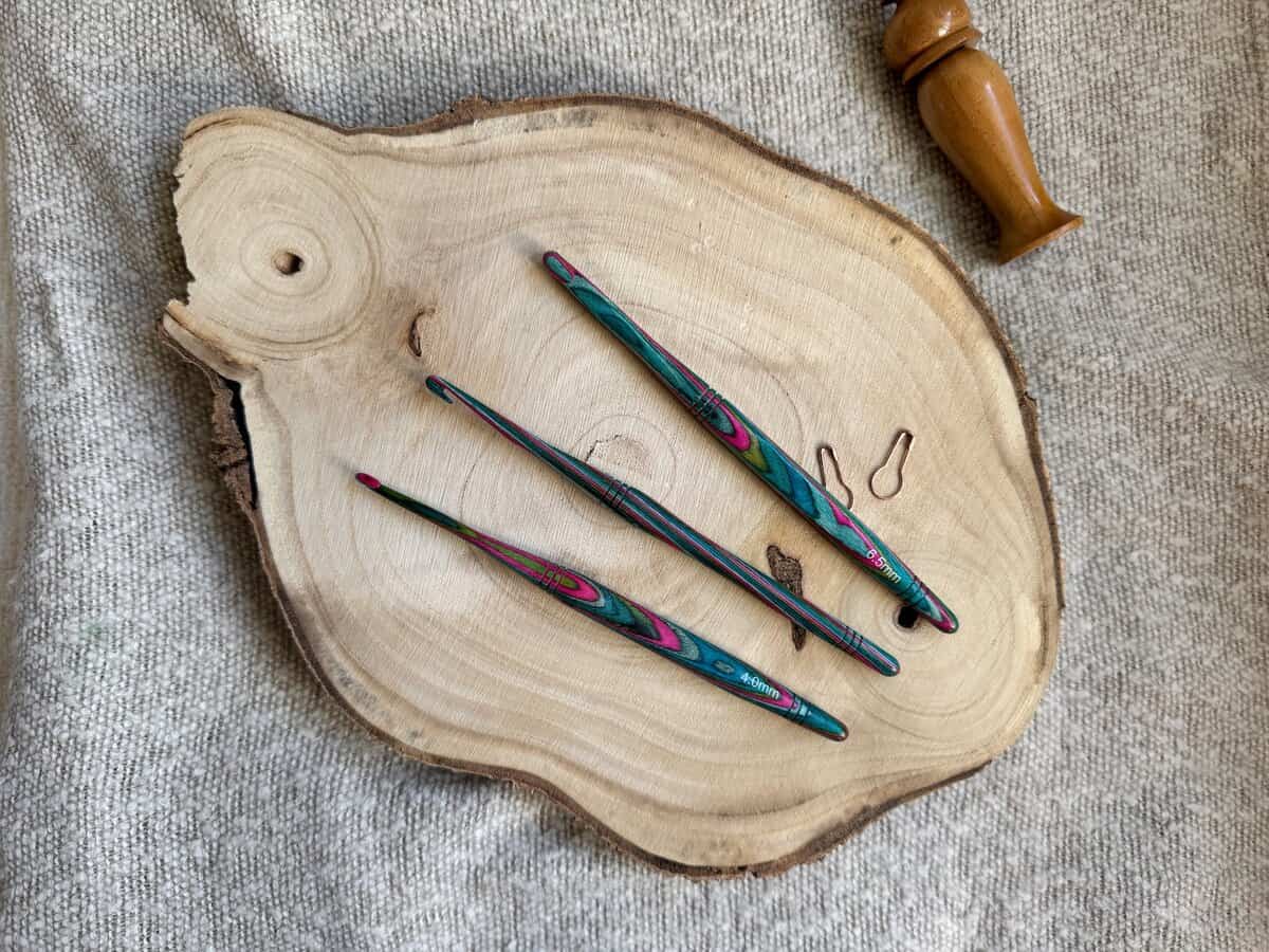 Three colorful crochet hooks in different crochet hook sizes are placed on a wooden slice, with a wooden handle tool and two stitch markers nearby on a textured fabric background.
