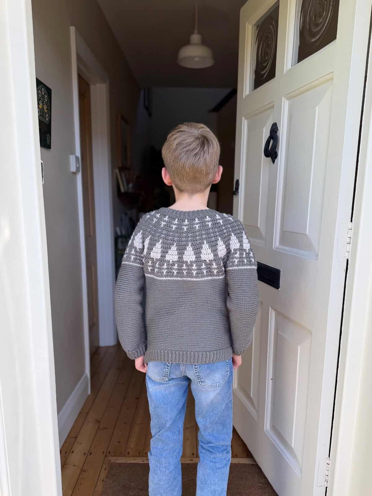 A young boy wearing a grey crochet Christmas sweater and blue jeans stands just inside an open white front door, facing away down a hallway.