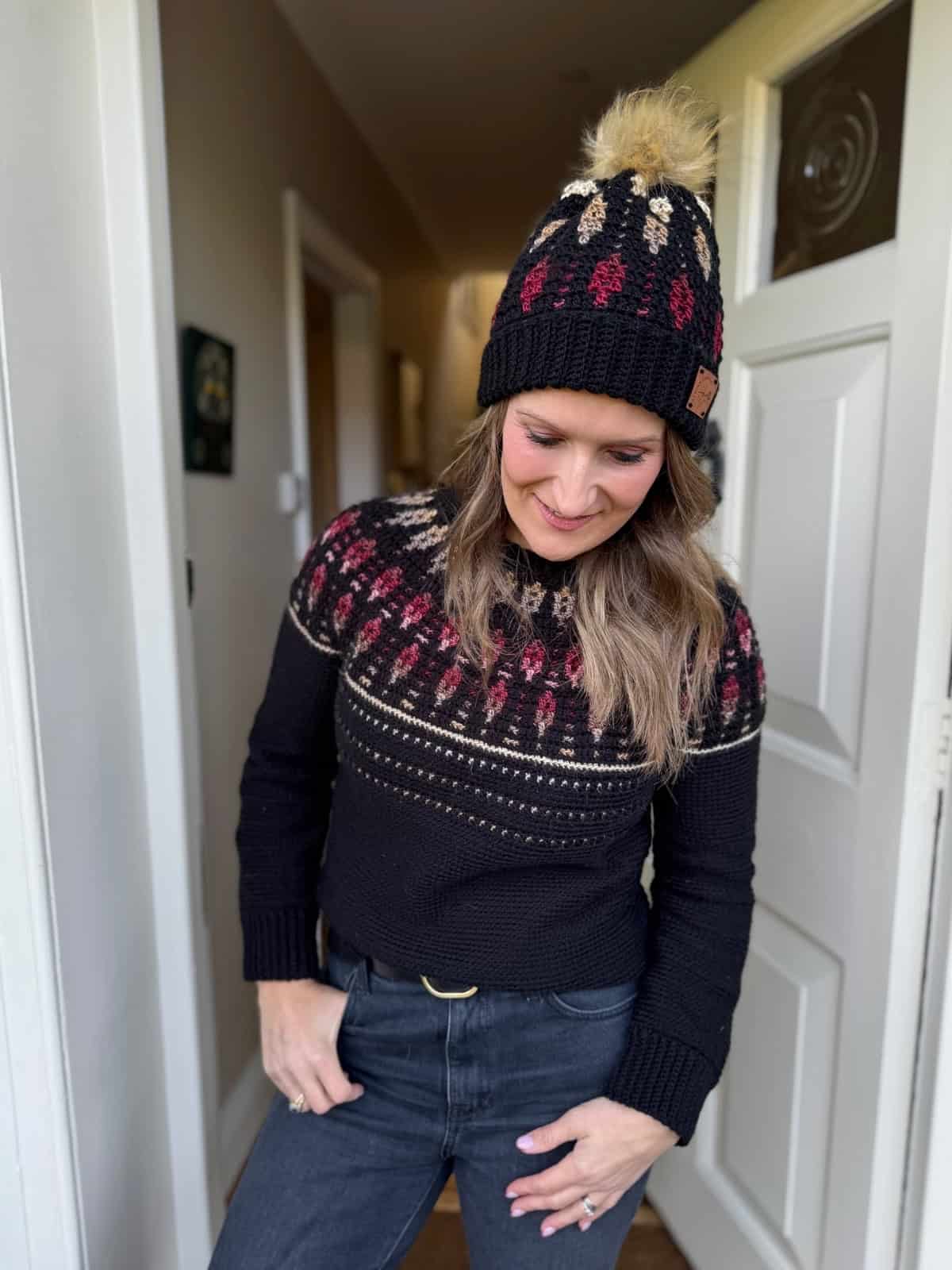 A woman stands indoors wearing a black crochet sweater with a patterned yoke and a matching black beanie with a pom-pom, looking down and smiling.