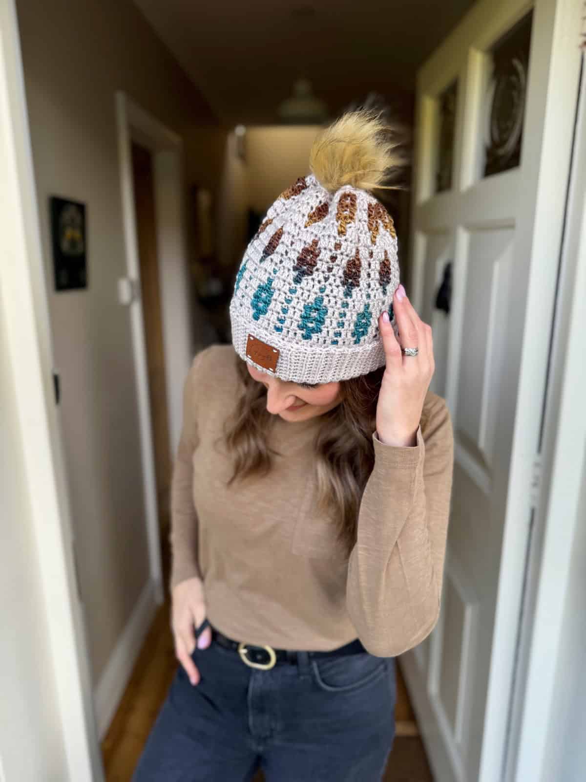 A woman wearing a mosaic crochet sweater pattern, black jeans, and a knit beanie with a pom-pom stands indoors, looking down and touching her hat.