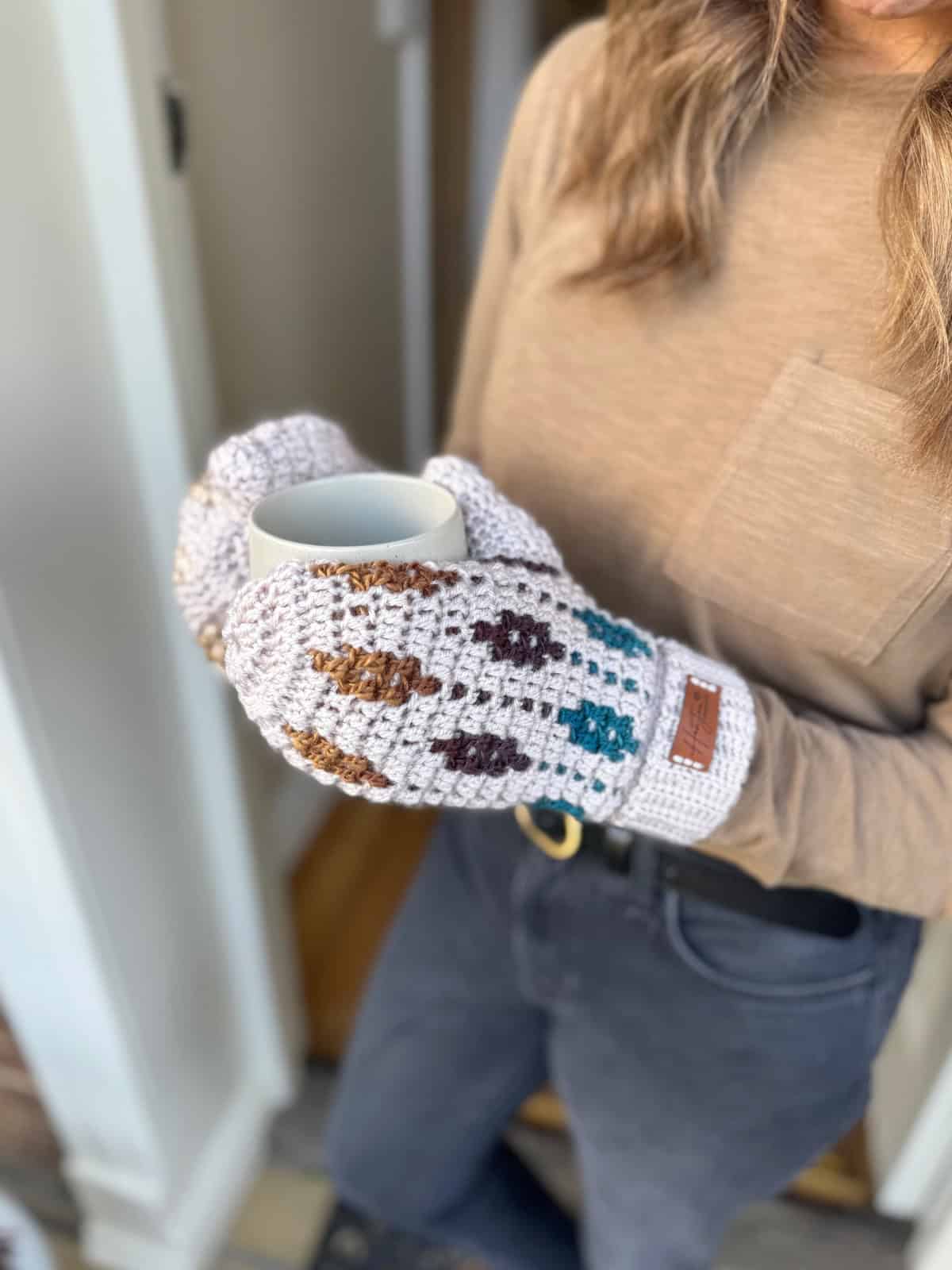 A person wearing patterned crochet mittens holds a white mug while standing indoors, dressed in a brown long-sleeve shirt, dark jeans, and a cozy mosaic crochet sweater pattern.