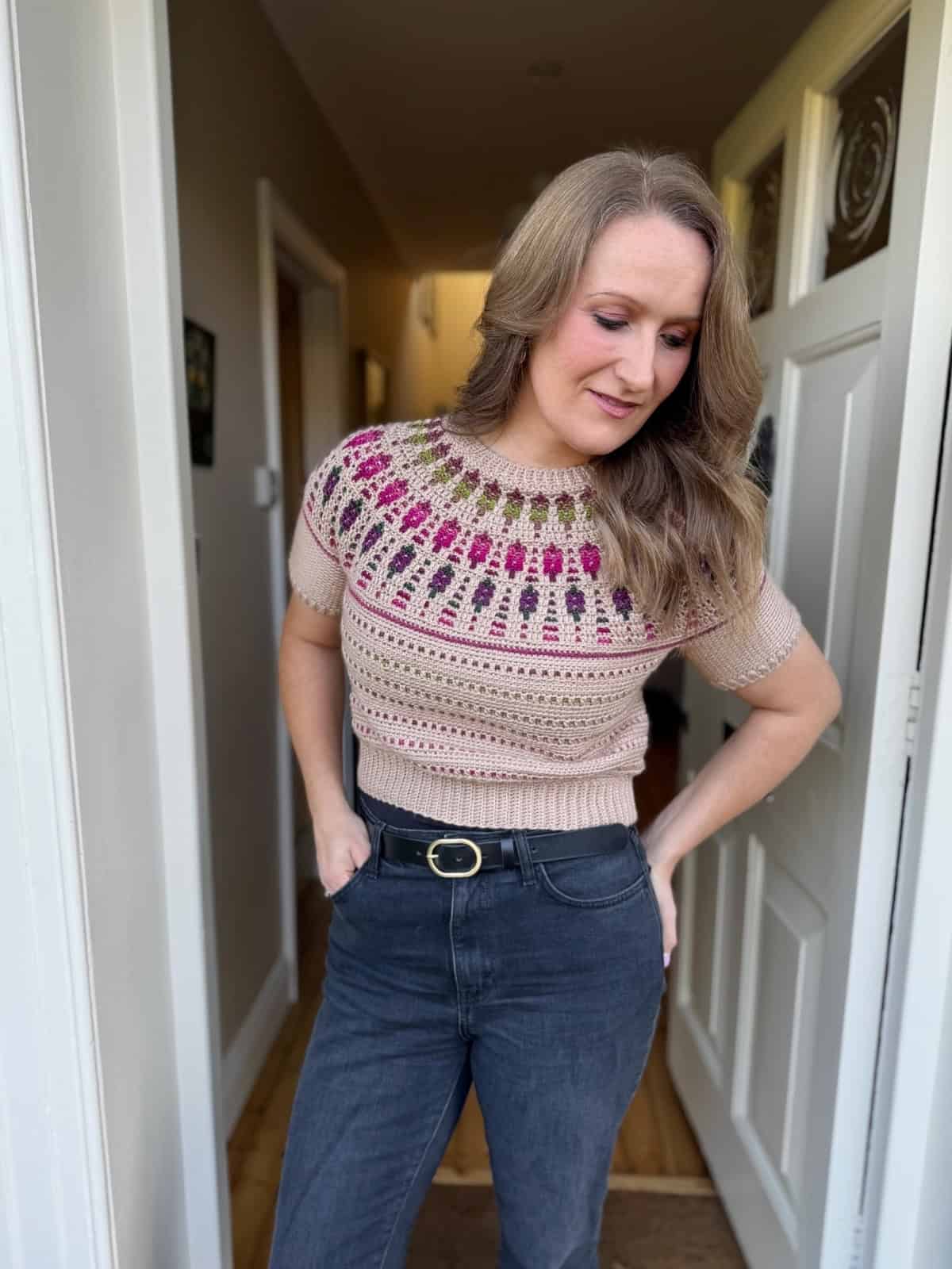 A woman stands indoors wearing a short-sleeved crochet sweater with a pink and purple patterned yoke, paired with high-waisted jeans and a black belt.