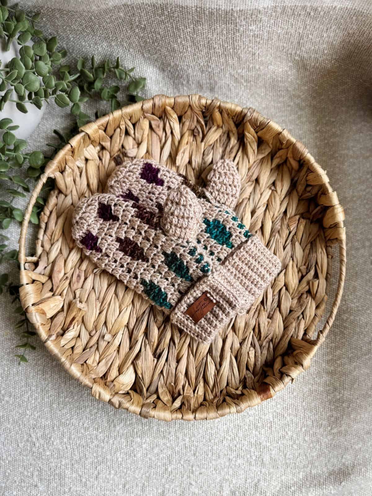 A pair of beige mosaic crochet mittens with green and purple accents is placed on a round woven tray, with green leaves partially visible on the side.