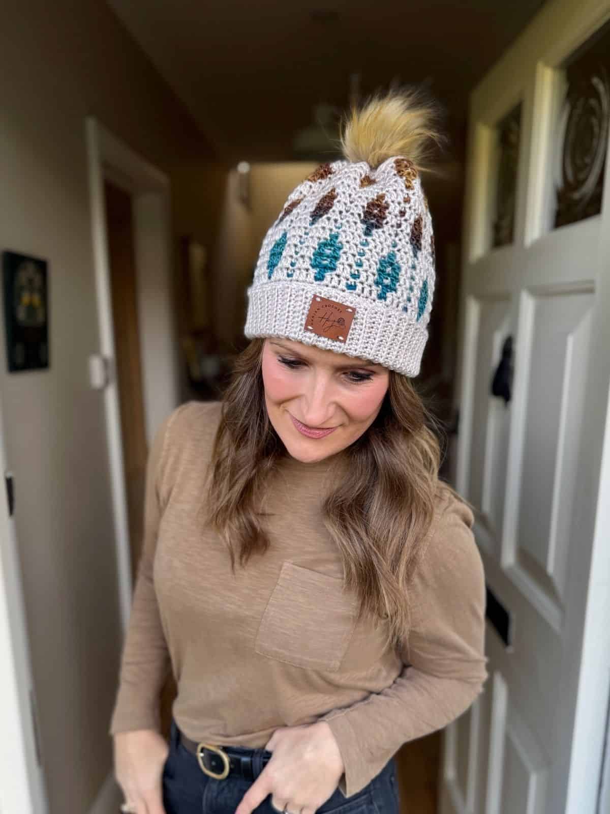 A woman wearing a tan long-sleeve shirt and a mosaic crochet beanie with a pom-pom stands indoors near a white door, looking down and smiling.