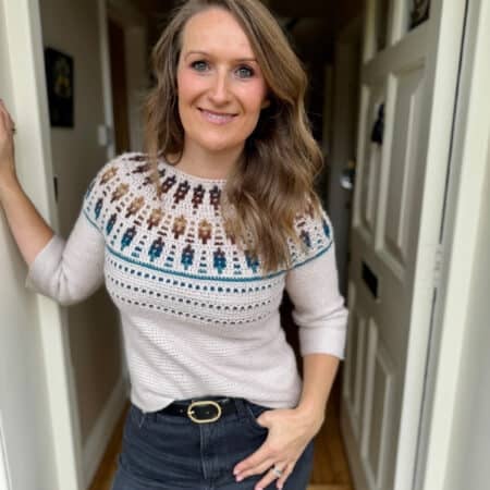 A woman with long, wavy hair stands in a doorway, wearing a mosaic crochet sweater pattern and dark jeans, smiling at the camera.
