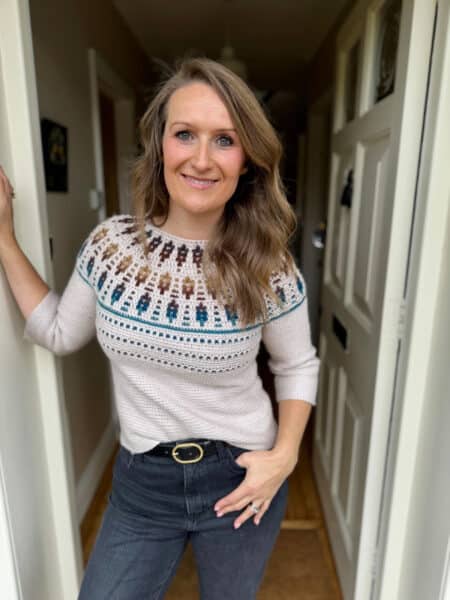 A woman with long, wavy hair stands in a doorway, wearing a mosaic crochet sweater pattern and dark jeans, smiling at the camera.