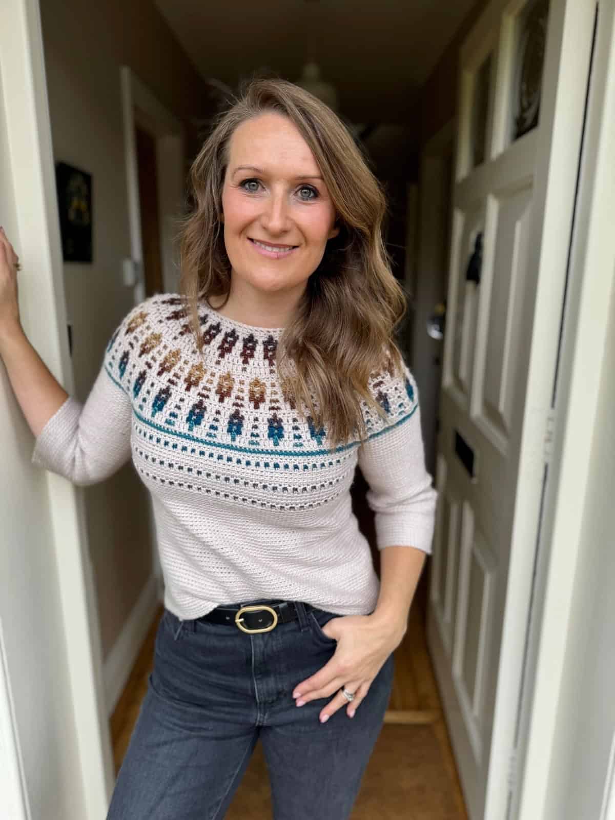 A woman with long, wavy hair stands in a doorway, wearing a mosaic crochet sweater pattern and dark jeans, smiling at the camera.