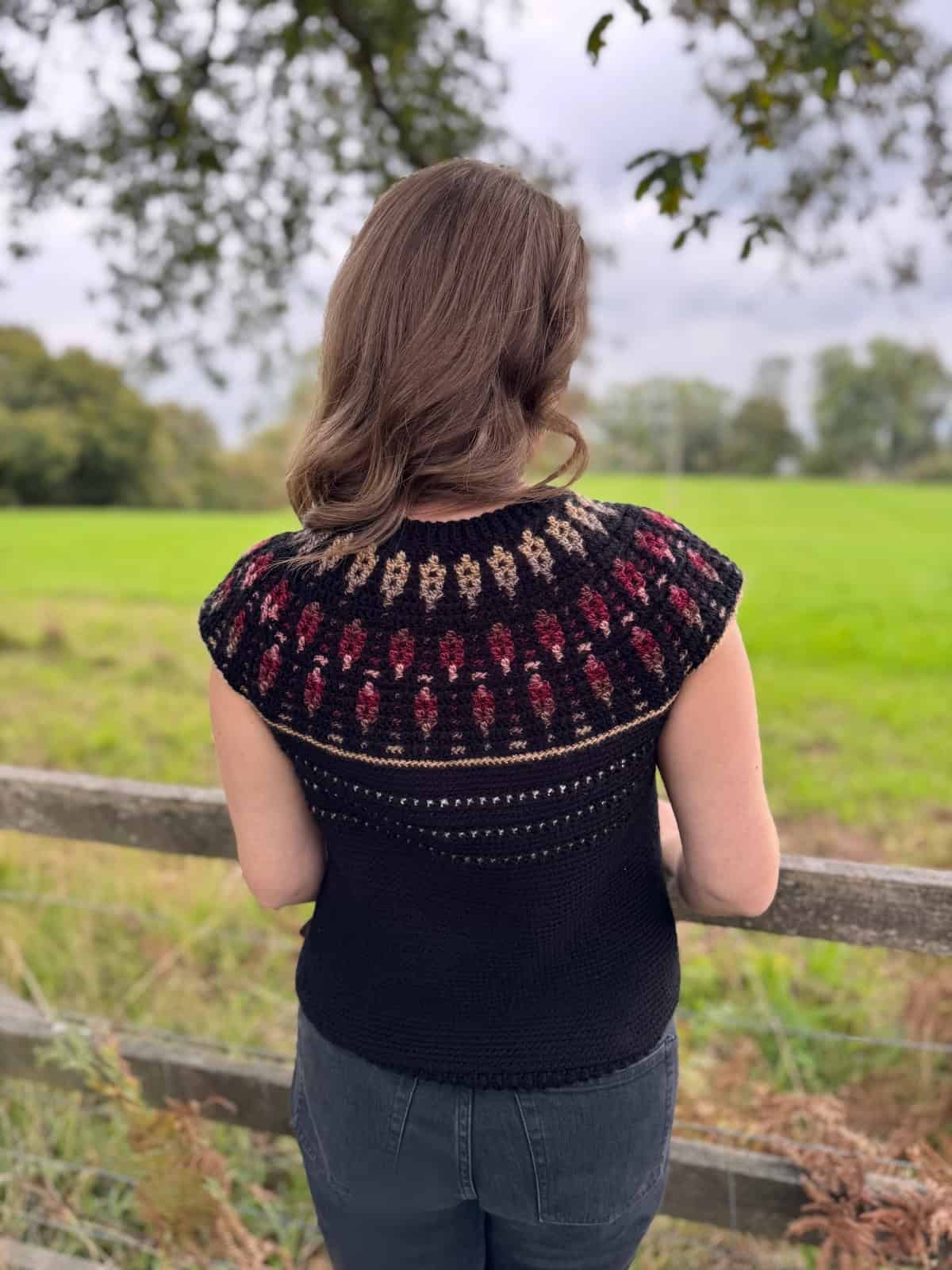 A person with shoulder-length brown hair faces a green field, wearing a black sleeveless knit top with a colorful yoke inspired by a mosaic crochet sweater pattern.