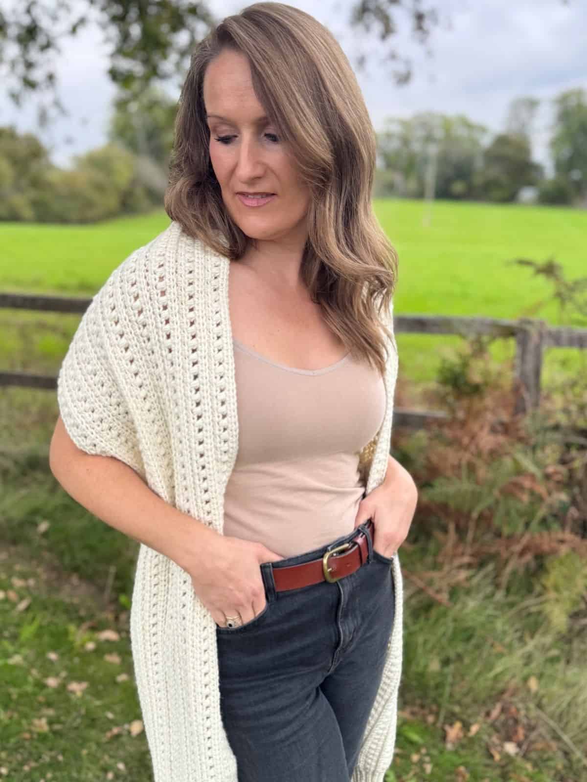 Woman standing outdoors in front of a field, wearing a beige tank top, black jeans with a brown belt, and a cream crochet sweater scarf—perfect inspiration if you're looking for a free pattern. She is looking down with hands in her pockets.