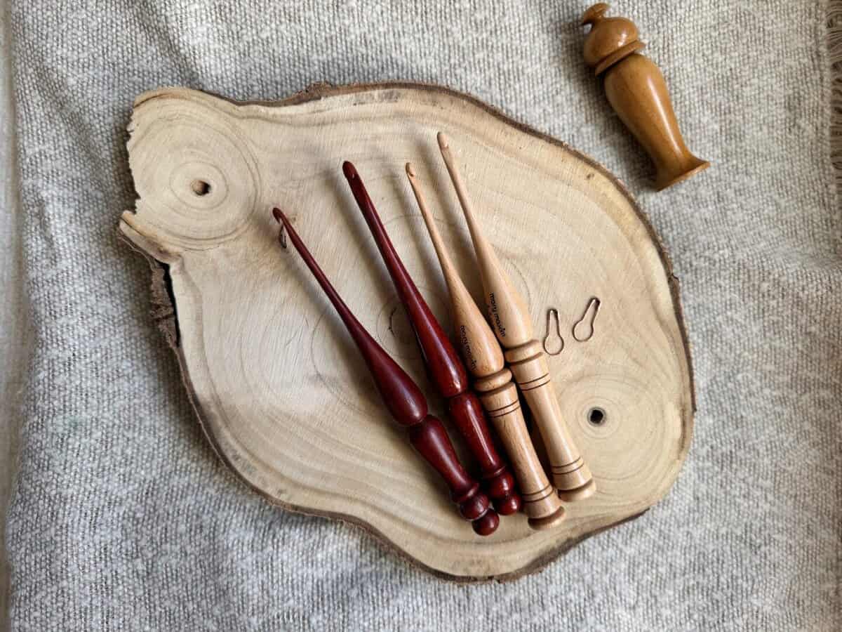 Five wooden crochet hooks in various crochet hook sizes and two stitch markers rest on a slice of wood placed on a light fabric background; a wooden closure lies nearby.