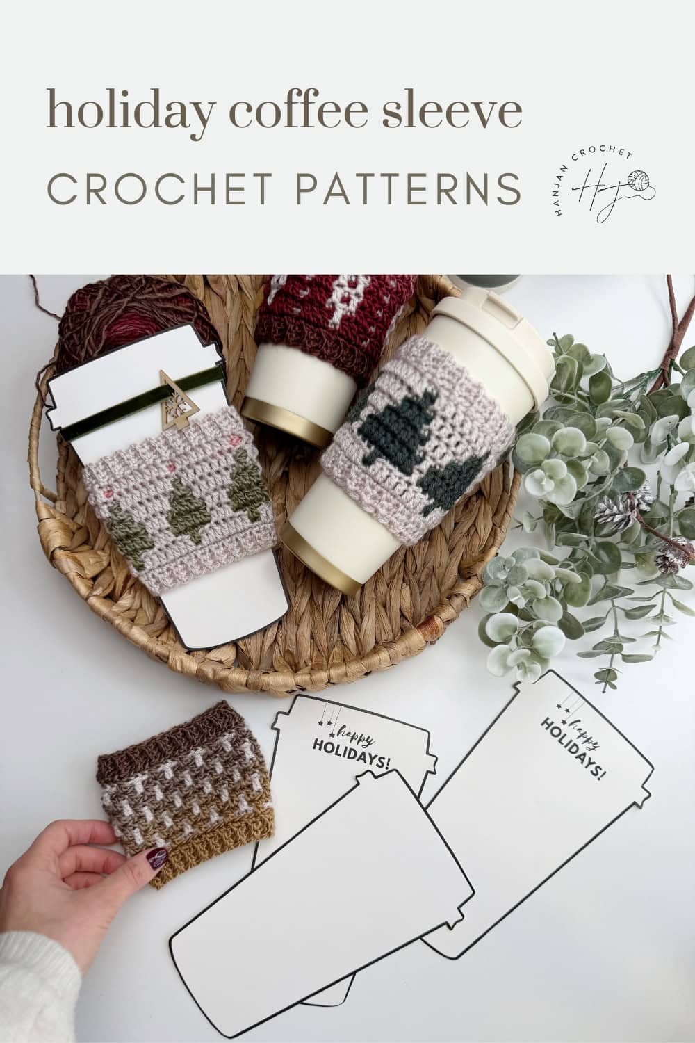 A wicker tray with crocheted holiday coffee sleeves on cups, yarn, greenery, and paper coffee cup templates labeled "Happy Holidays," featuring a festive Crochet Coffee Sleeve Pattern.