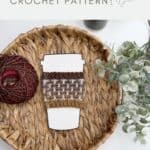 A crochet coffee cup cozy displayed on a paper cup cutout, placed on a woven tray with a ball of yarn and greenery. Text above reads "coffee cup cozy CROCHET PATTERN." Perfect Crochet Coffee Sleeve Pattern for your favorite mug!.